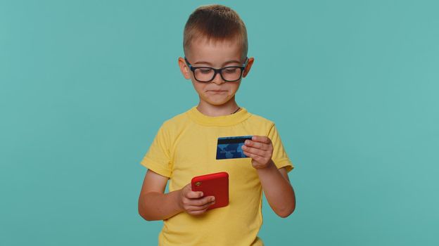 Little toddler children boy in yellow t-shirt using credit bank card and smartphone while transferring money, purchases online shopping. Young preschool kid isolated alone on blue studio background
