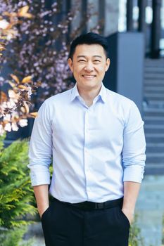 Vertical frame, Asian man in white shirt looks at camera and smiles, hands in pockets