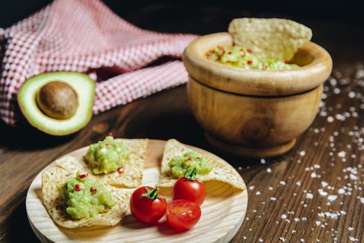 homemade guacamole in a wood bowl with nachos next to an avocado and a kitchen rag, typical mexican healthy vegan cuisine with rustic dark food photo style