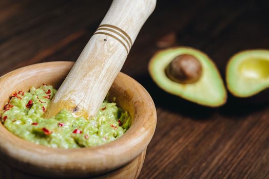 traditional guacamole in a wood bowl and cut half avocado on wooden table, typical mexican healthy vegan cuisine with rustic dark food photo style, selective focus
