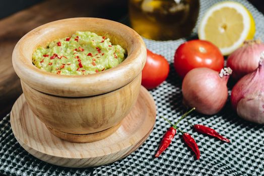 fresh guacamole in a wood bowl next to ingredients, typical mexican healthy vegan cuisine with rustic dark food photo style