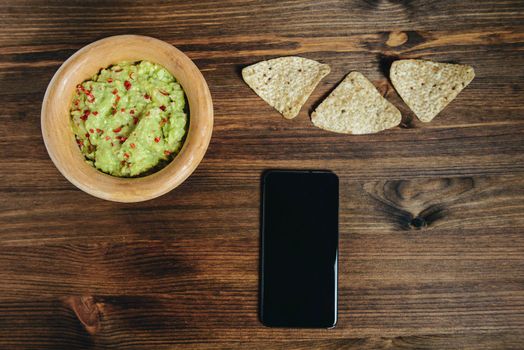 homemade guacamole with nachos and a phone in a wood bowl on a wooden table, typical mexican healthy vegan cuisine, top view and copy space for text