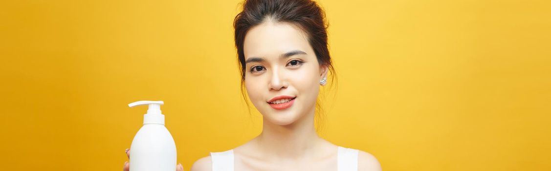 Smiling young asian woman showing skincare product on yellow background