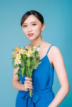 Attractive Asian woman in blue dress holding bouquet of flowers over blue background