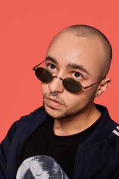 Close-up studio shot of a good-looking tattoed bald male in sunglasses, with a pirsing ring in his nose, wearing black trendy t-shirt with print and sport suit, looking up and posing against a pink background with copy space. People, style and fashion concept. 90s style