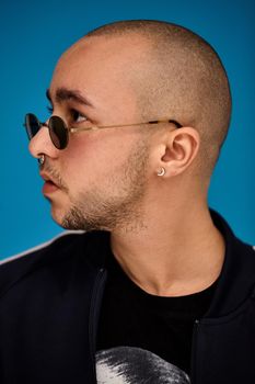 Close-up studio shot of a handsome tattoed bald person in sunglasses, with a pirsing ring in his nose, wearing black trendy t-shirt with print and sport suit, posing sideways against a blue background with copy space. People, style and fashion concept. 90s style