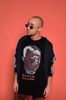 Close-up studio shot of a young tattoed bald male in sunglasses, with a pirsing ring in his nose, wearing black trendy t-shirt with print and sport suit, looking away while posing against a pink background with copy space. People, style and fashion concept. 90s style