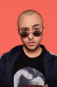 Close-up studio shot of a good-looking tattoed bald man in sunglasses, with a pirsing ring in his nose, wearing black trendy t-shirt with print and sport suit, posing against a pink background with copy space. People, style and fashion concept. 90s style