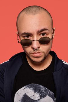 Close-up studio shot of a handsome tattoed bald person in sunglasses, with a pirsing ring in his nose, wearing black trendy t-shirt with print and sport suit, looking at you while posing against a pink background with copy space. People, style and fashion concept. 90s style