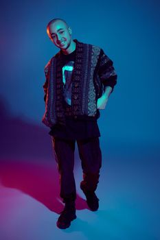Full-length studio shot of a handsome tattoed bald person with a pirsing ring in his nose, wearing dark jacket, black trendy t-shirt with print, sneakers and sweatpants, looking at the camera and smiling while posing against a colorful background with copy space. People, style and fashion concept. 90s style