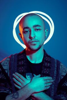 Close-up studio shot of a enjoying tattoed bald person with a pirsing ring in his nose, wearing dark jacket, black trendy t-shirt with print, crossed his arms and posing against a blue background with copy space. People, style and fashion concept. 90s style