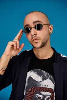 Close-up studio shot of a young tattoed bald fellow in sunglasses, with a pirsing ring in his nose, wearing black trendy t-shirt with print and sport suit, looking at the camera and posing against a blue background with copy space. People, style and fashion concept. 90s style