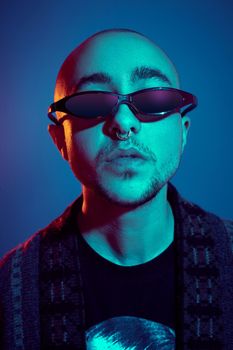 Close-up studio shot of a relaxed tattoed bald guy in sunglasses, with a pirsing ring in his nose, wearing dark jacket, black trendy t-shirt with print, posing against a blue background with copy space. People, style and fashion concept. 90s style