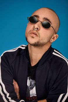 Close-up studio shot of a good-looking tattoed bald fellow in sunglasses, with a pirsing ring in his nose, wearing black trendy t-shirt with print and sport suit, posing against a blue background with copy space. People, style and fashion concept. 90s style