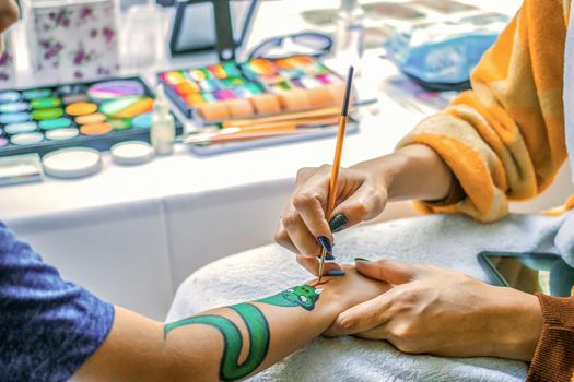 Young girl artist draws a dragon on child's hand. Painting skin with paints, close-up, without faces.