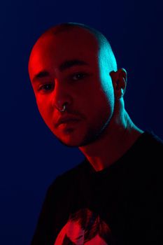 Close-up studio shot of a good-looking tattoed bald person with a pirsing ring in his nose, wearing black trendy t-shirt with print, posing and looking at the camera against a blue background with copy space. People, style and fashion concept. 90s style