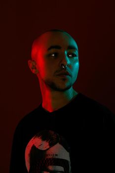 Close-up studio shot of a good-looking tattoed bald man with a pirsing ring in his nose, wearing black trendy t-shirt with print, posing and looking away against a red background with copy space. People, style and fashion concept. 90s style