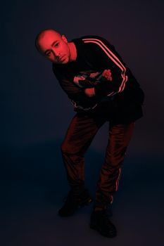 Full-length studio shot of a handsome tattoed bald person with a pirsing ring in his nose, wearing black trendy t-shirt with print, sport suit and sneakers, looking at the camera while posing with crossed hands against a dark blue background with copy space. People, style and fashion concept. 90s style