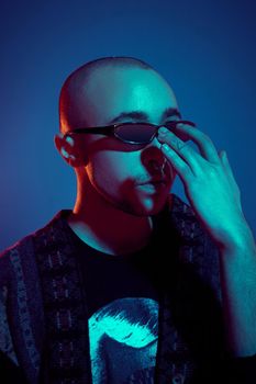 Close-up studio shot of a enjoying tattoed bald guy is straightening sunglasses, with a pirsing ring in his nose, wearing dark jacket, black trendy t-shirt with print, posing against a blue background with copy space. People, style and fashion concept. 90s style