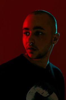 Close-up studio shot of a good-looking tattoed bald male with a pirsing ring in his nose, wearing black trendy t-shirt with print, posing against a red background with copy space. People, style and fashion concept. 90s style