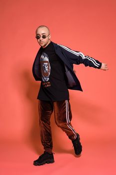 Full-length studio shot of a nice tattoed bald person in sunglasses, with a pirsing ring in his nose, wearing black trendy t-shirt with print, sport suit and sneakers, spinning and posing against a pink background with copy space. People, style and fashion concept. 90s style