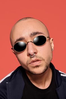 Close-up studio shot of a handsome tattoed bald man in sunglasses, with a pirsing ring in his nose, wearing black trendy t-shirt with print and sport suit, looking at the camera while posing against a pink background with copy space. People, style and fashion concept. 90s style