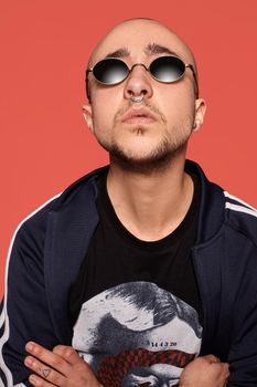 Close-up studio shot of a relaxed tattoed bald person in sunglasses, with a pirsing ring in his nose, wearing black trendy t-shirt with print and sport suit, looking up while posing against a pink background with copy space. People, style and fashion concept. 90s style