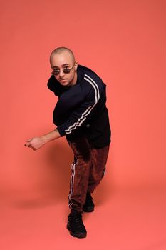 Full-length studio shot of a young tattoed bald fellow in sunglasses, with a pirsing ring in his nose, wearing black trendy t-shirt with print, sport suit and sneakers, fooling around while posing against a pink background with copy space. People, style and fashion concept. 90s style