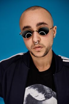 Close-up studio shot of a relaxed tattoed bald male in sunglasses, with a pirsing ring in his nose, wearing black trendy t-shirt with print and sport suit, looking at the camera while posing against a blue background with copy space. People, style and fashion concept. 90s style