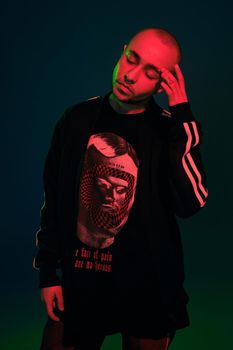 Close-up studio shot of a adult tattoed bald guy with a pirsing ring in his nose, wearing black trendy t-shirt with print and sport suit, closed his eyes while posing against a colorful background with copy space. People, style and fashion concept. 90s style