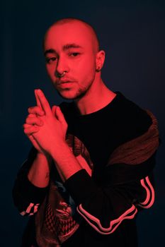 Close-up studio shot of a good-looking tattoed bald person with a pirsing ring in his nose, wearing black trendy t-shirt with print and sport suit, folded his arms like a gun posing against a dark blue background with copy space. People, style and fashion concept. 90s style