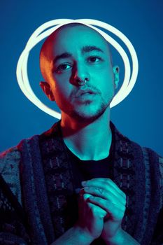 Close-up studio shot of a enjoying tattoed bald guy with a pirsing ring in his nose, wearing dark jacket, black trendy t-shirt with print, praying while posing against a blue background with copy space. People, style and fashion concept. 90s style