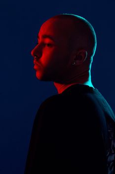 Close-up studio shot of a relaxed tattoed bald male with a pirsing ring in his nose, wearing black trendy t-shirt with print, posing sideways against a blue background with copy space. People, style and fashion concept. 90s style