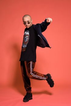 Full-length studio shot of a handsome tattoed bald man in sunglasses, with a pirsing ring in his nose, wearing black trendy t-shirt with print, sport suit and sneakers, raised his leg while posing against a pink background with copy space. People, style and fashion concept. 90s style