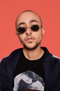 Close-up studio shot of a good-looking tattoed bald fellow in sunglasses, with a pirsing ring in his nose, wearing black trendy t-shirt with print and sport suit, looking straight and posing against a pink background with copy space. People, style and fashion concept. 90s style