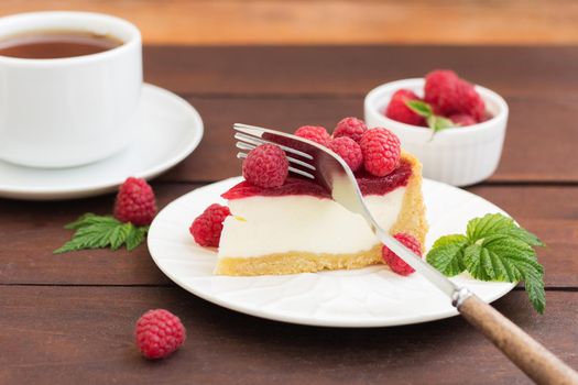 A fork cuts a piece of raspberry pie (cheesecake) lying on a plate, on a wooden background