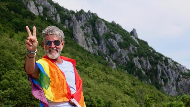 Portrait of a gray-haired senior elderly Caucasian man bisexuality with a beard and sunglasses with a rainbow LGBTQIA peace flag in mountains. Celebrates Pride Month, Rainbow Flag Day, gay parade