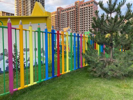 Colorful fence of a kindergarten or park in the form of colored pencils standing in a row. Beautiful funny kindergarten fence and a multi-storey house under construction in the background. New neighborhood, new building concept
