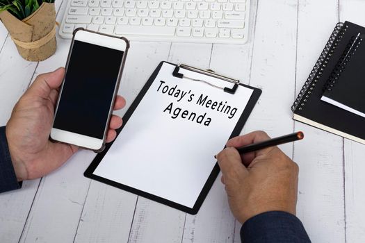 Hand with pencil writing Today's Meeting Agenda on the black clipboard with holding a smartphone . Business and education concept.