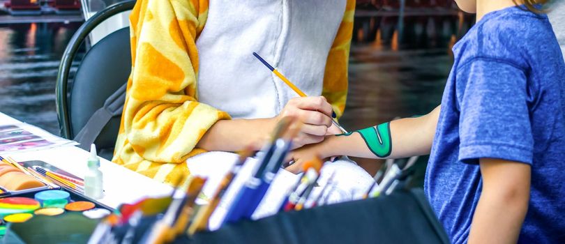 Young girl artist draws a dragon on child's hand. Process of drawing on skin, close-up, banner.