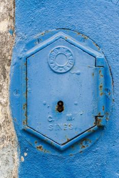 Old rusty blue painted iron lid of a valve box of the SERVICO DE AGUA PUBLIC WATER SUPPLY on the chipped white - blue painted wall of a house in the Old Town area, Portugal