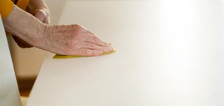 An elderly woman is doing cleaning in her cozy home, in the kitchen she holds a rag in her hand and removes the crumbs from the white table with detergent, close-up view.