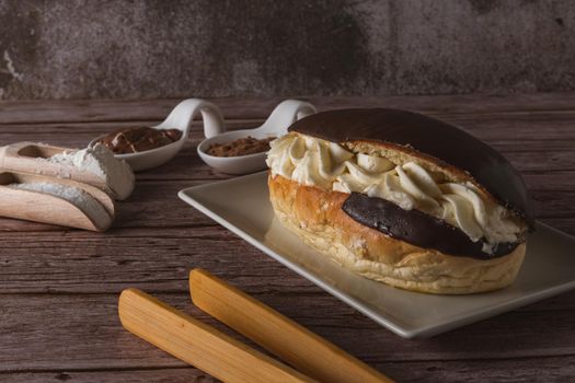 chocolate-covered cream-filled bun on a white plate with the ingredients and wooden tongs.