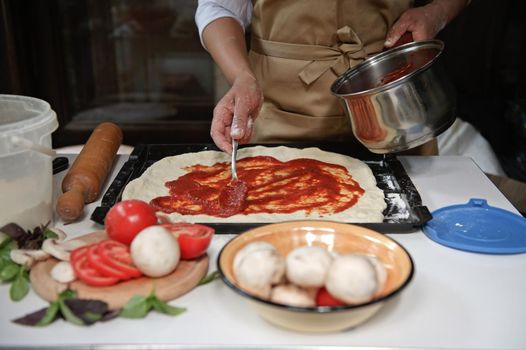 Front view. Close-up housewife in beige chef apron spreading freshly made tomato sauce on rolled out dough in pizza pan. Fresh organic ingredients on kitchen table. Cooking. Culinary. Italian cuisine