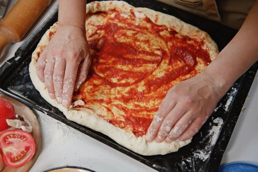 Close-up chef pizzaiolo makes homemade Italian pizza with tomato sauce, fresh mushrooms, feta cheese and basil leaves in a rustic kitchen. Traditional Italian cuisine. Healthy eating. Food consumerism