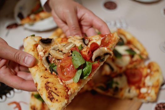 Selective focus, woman hands holding a slice of delicious appetizing homemade freshly baked Italian pizza with organic juicy tomatoes, mushroom champignons, mozzarella cheese and fragrant basil leaves