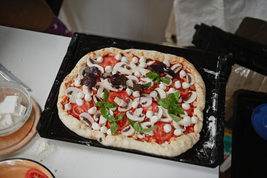 Yummy raw Italian vegetarian pizza with fresh organic ingredients: sliced champignons, ripe juicy tomatoes, mozzarella cheese and feta in the sides on a pizza pan, on white table background. Top view