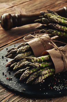 Bunches of an edible, seasonal stems of asparagus on a stone slate, wooden background. Fresh, green vegetables with seasonings, kitchen pestle. Healthy eating. Fall harvest, agricultural farming concept.