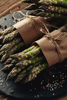 Bunches of an edible, organic spears of asparagus on a stone slate, wooden background. Fresh, green vegetables with seasonings, top view. Healthy eating. Fall harvest, agricultural farming concept.