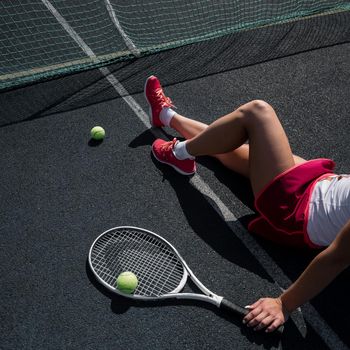 A faceless girl in a sports skirt sits on a tennis court and holds a rocket. Top view of female legs
